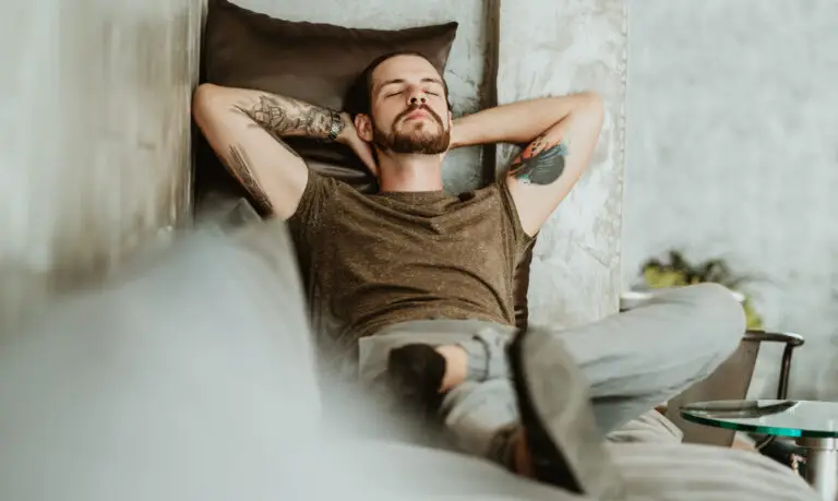 Young, bearded man resting on a cozy modern sofa in a light-colored living room.