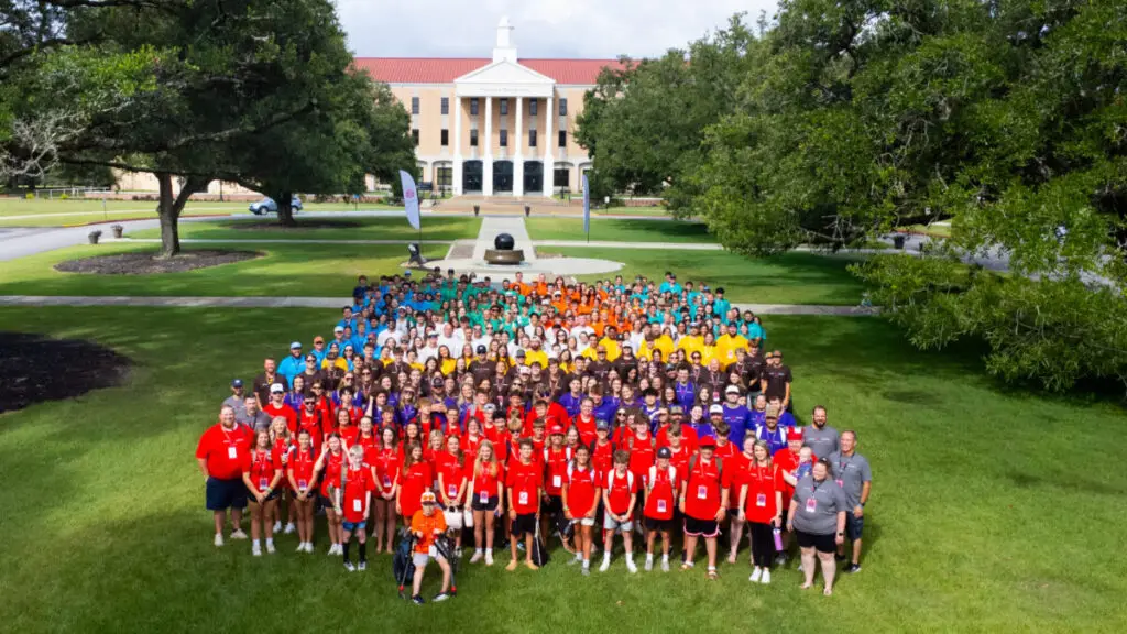 A large group of students standing in groups with matching colored shirts on the lawn of the University of Mobile during Super Summer Alabama.