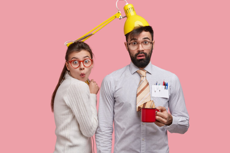 A bearded man with dark hair wearing glasses is seen wearing a yellow desk lamp on his head. He is holding a red mug. To his left, a woman with brown hair tied back and wearing glasses and a white sweater looks surprised. The background is pink.