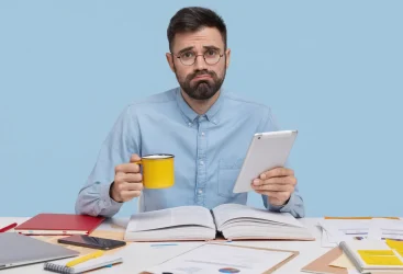 A man with a beard wearing glasses and a blue collared shirt sits at a desk looking directly at the camera with a sad expression. He's holding a yellow mug in one hand and a tablet in the other. On the desk is an open book, notebooks, papers, a cellphone, and a laptop. The setting is an indoor workplace with a light blue background.