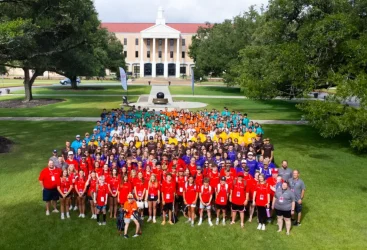 A large group of students standing in groups with matching colored shirts on the lawn of the University of Mobile during Super Summer Alabama.