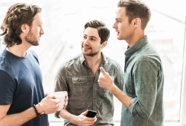 Three men standing talking, one with a cup of coffee, one with a smartphone.